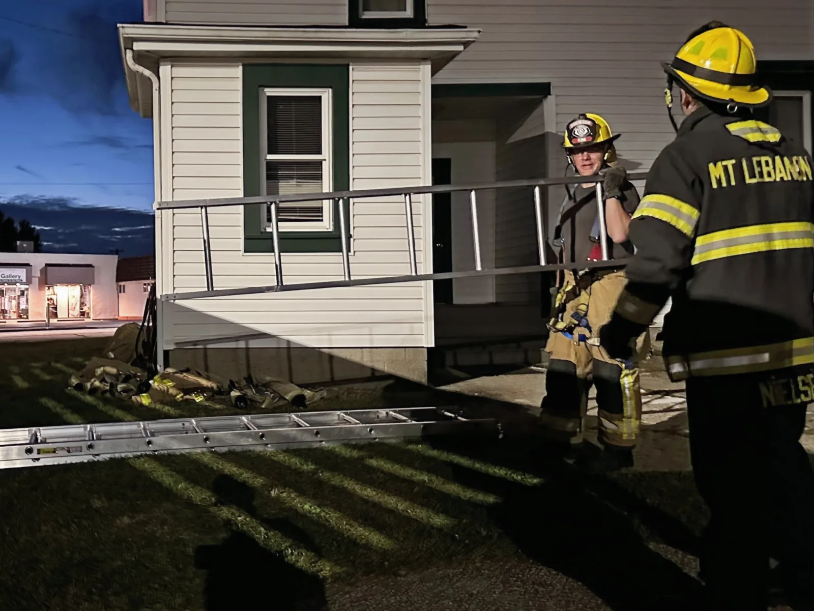 Mt. Lebanon firefighters working outside a house at dusk, a ladder on the ground.