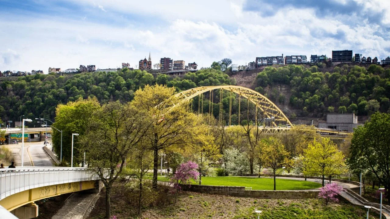 A tied-arch bridge over the river below a Pittsburgh hillside.