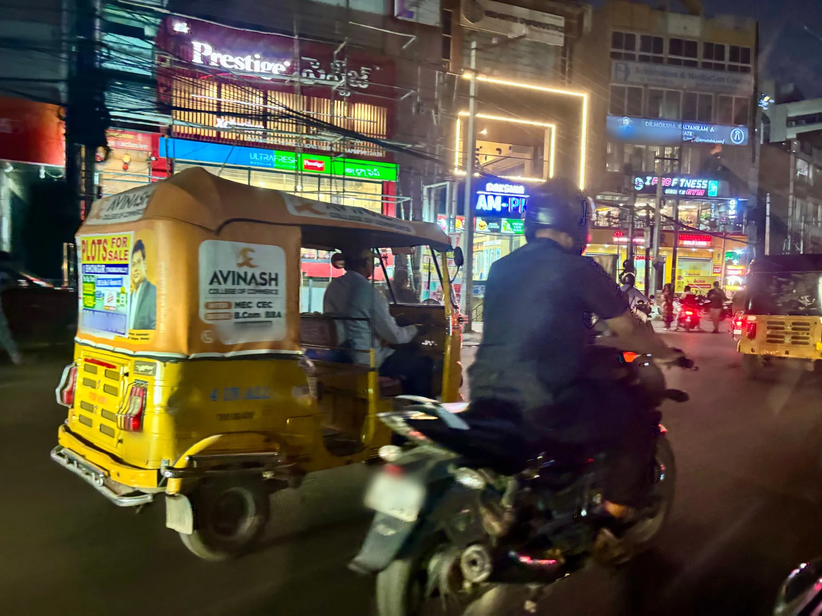 A yellow auto rickshaw wrapped in advertisements for land plots and a college of commerce moves through evening traffic in Hyderabad beside a motorcyclist, with lit shopfronts behind.