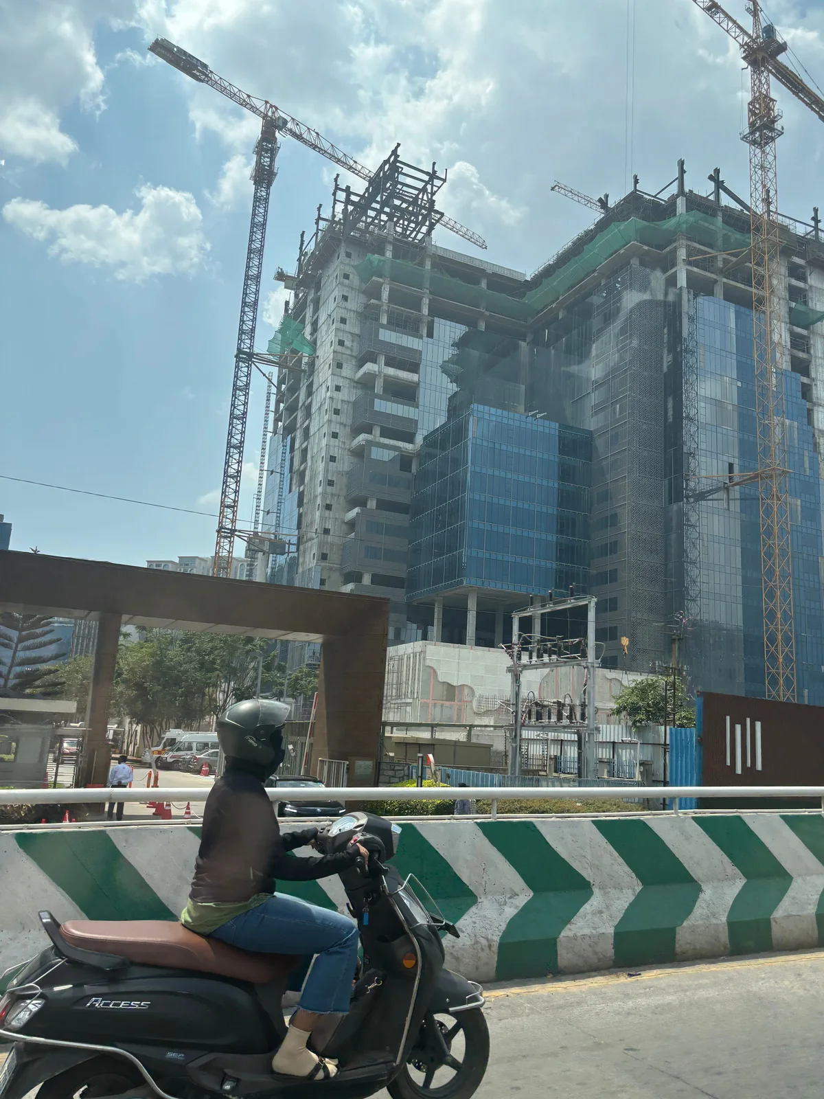 Two tower cranes stand over a glass and concrete high-rise under construction in Hyderabad while a woman on a scooter passes a green and white striped road barrier in the foreground.